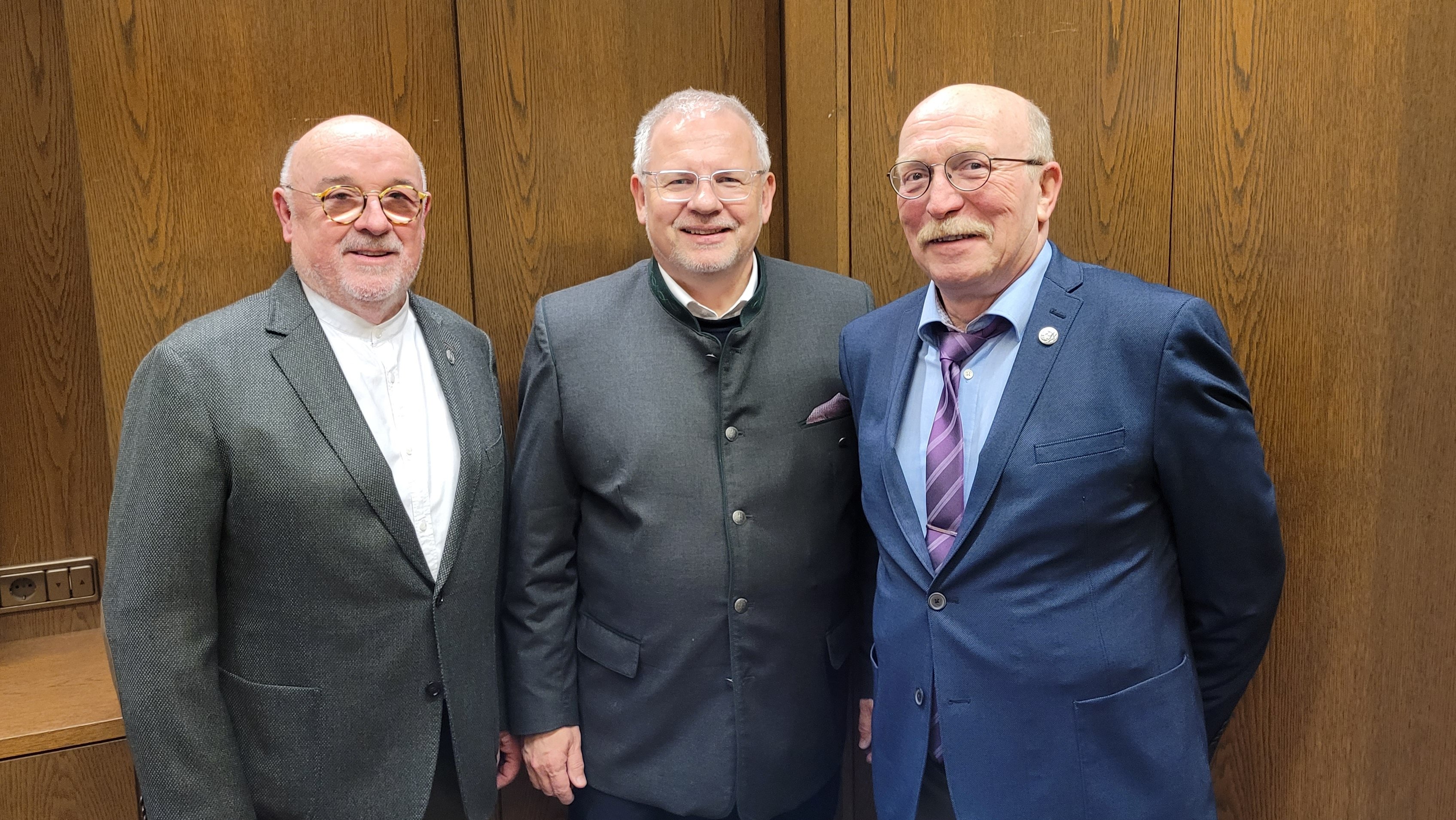 Drei Männer in Anzügen stehen vor einer mit dunklem Holz verkleideten Wand.