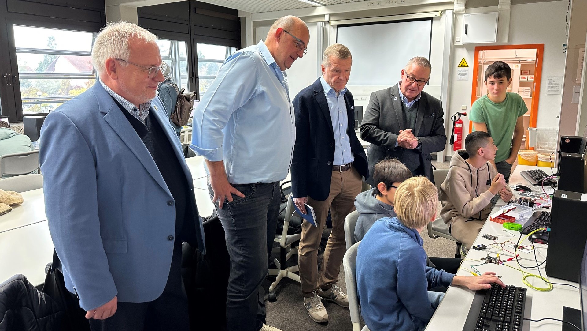 Landrat Hans-Ulrich Ihlenfeld, Lehrer Thomas Ehrmann, Gerhard Bugla, Rainer Bade stehen in einem Klassenraum. Vor ihnen sitzen drei Schüler vor Computern. Ein weiterer Schüler steht dabei.