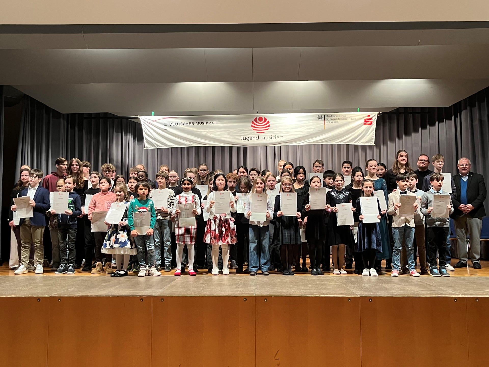 Eine Gruppe von etwa 100 Kindern steht auf einer Bühne und hält Urkunden hoch.