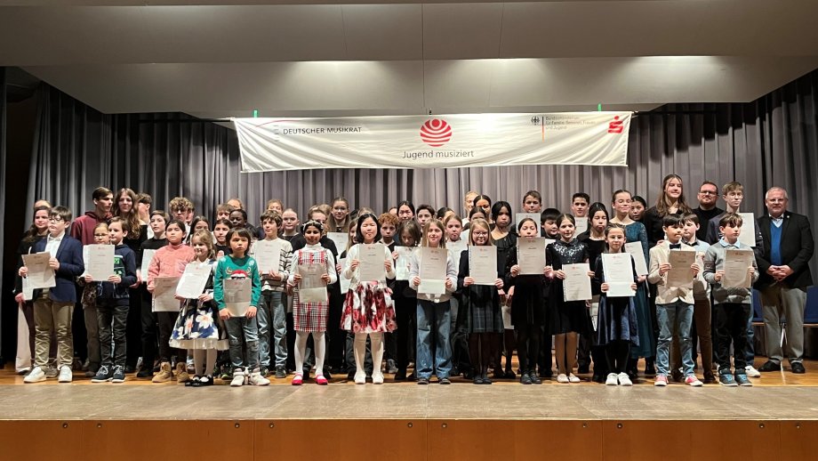 Eine Gruppe von etwa 100 Kindern steht auf einer Bühne und hält Urkunden hoch.