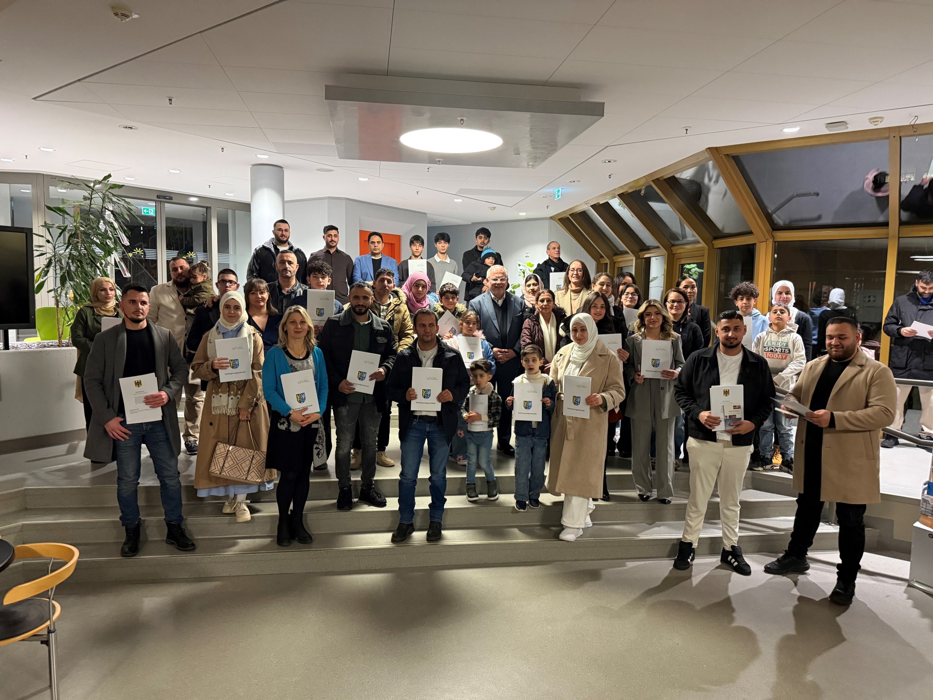 Eine Gruppe von Menschen hält ihre Urkunden hoch. Es sind 52 Personen, die neu eingebürgert wurden. In der Mitte steht Landrat Hans-Ulrich Ihlenfeld.