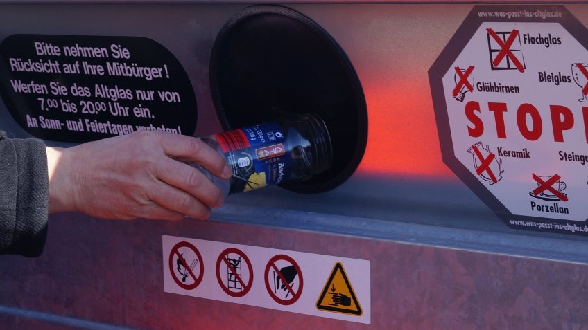 Eine Hand hält eine Flasche in die Öffnung eines Altglascontainers.