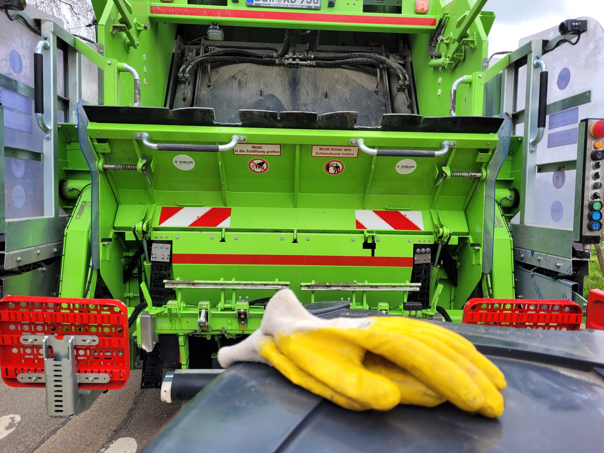 Gelbe Arbeitshandschuhe auf dem Deckel einer Mülltonne. Dahinter ein Müllsammelfahrzeug.