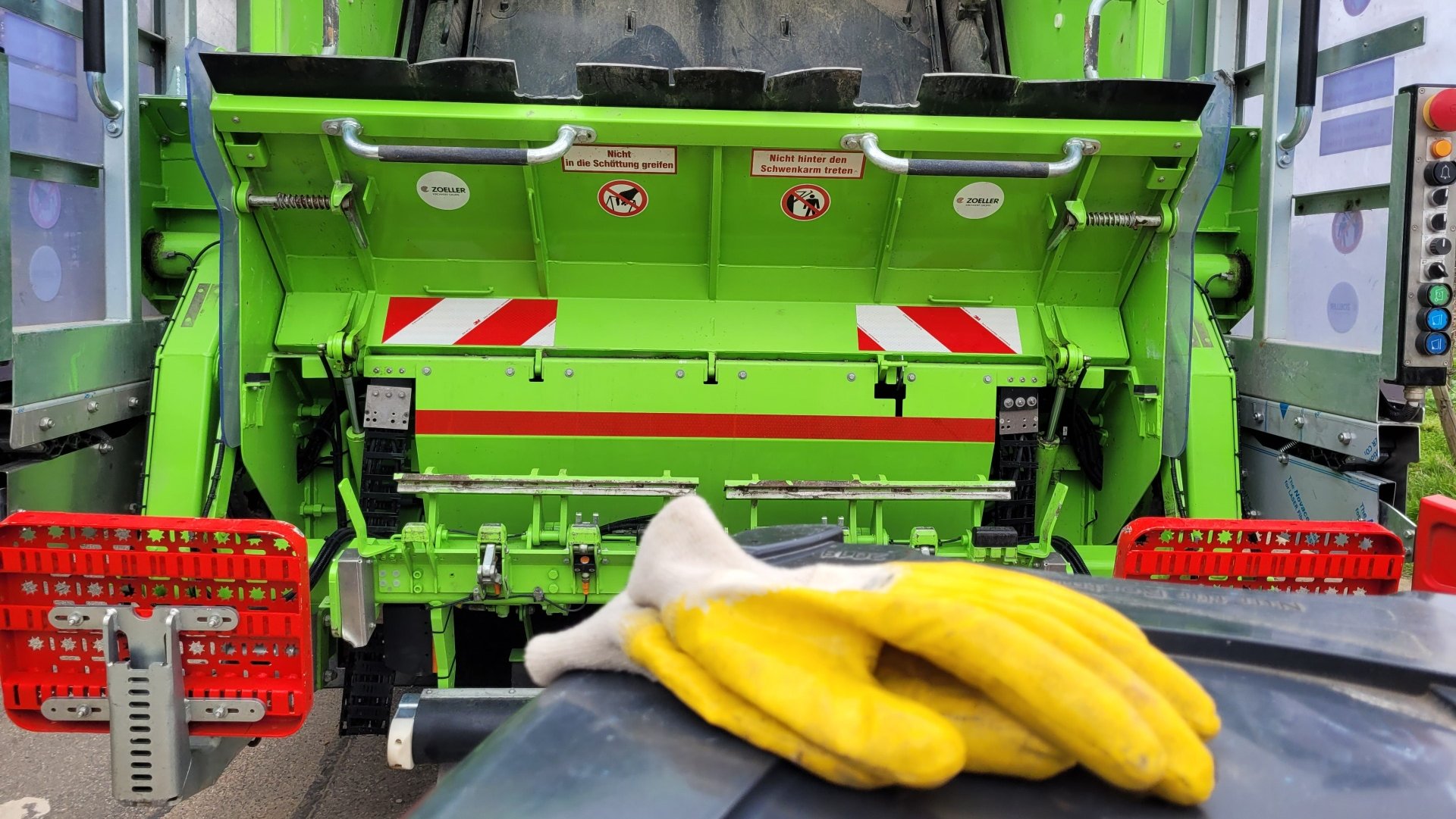 Gelbe Arbeitshandschuhe auf dem Deckel einer Mülltonne. Dahinter ein Müllsammelfahrzeug.