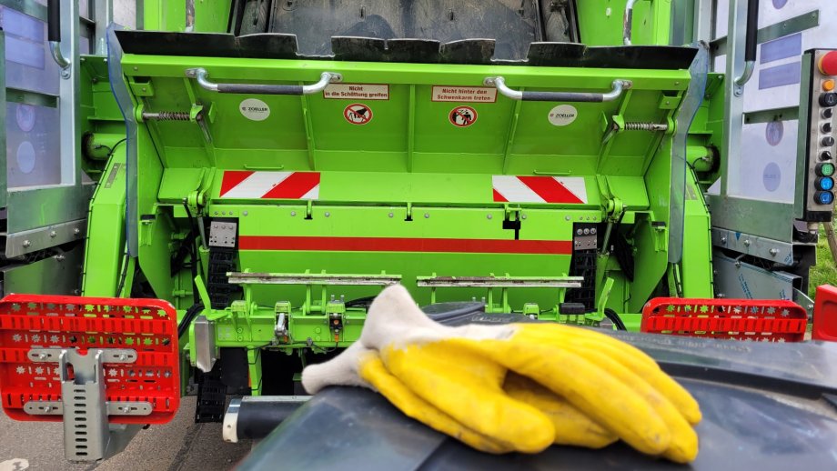 Gelbe Arbeitshandschuhe auf dem Deckel einer Mülltonne. Dahinter ein Müllsammelfahrzeug.