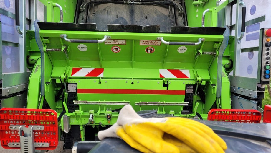 Gelbe Arbeitshandschuhe auf dem Deckel einer Mülltonne. Dahinter ein Müllsammelfahrzeug.