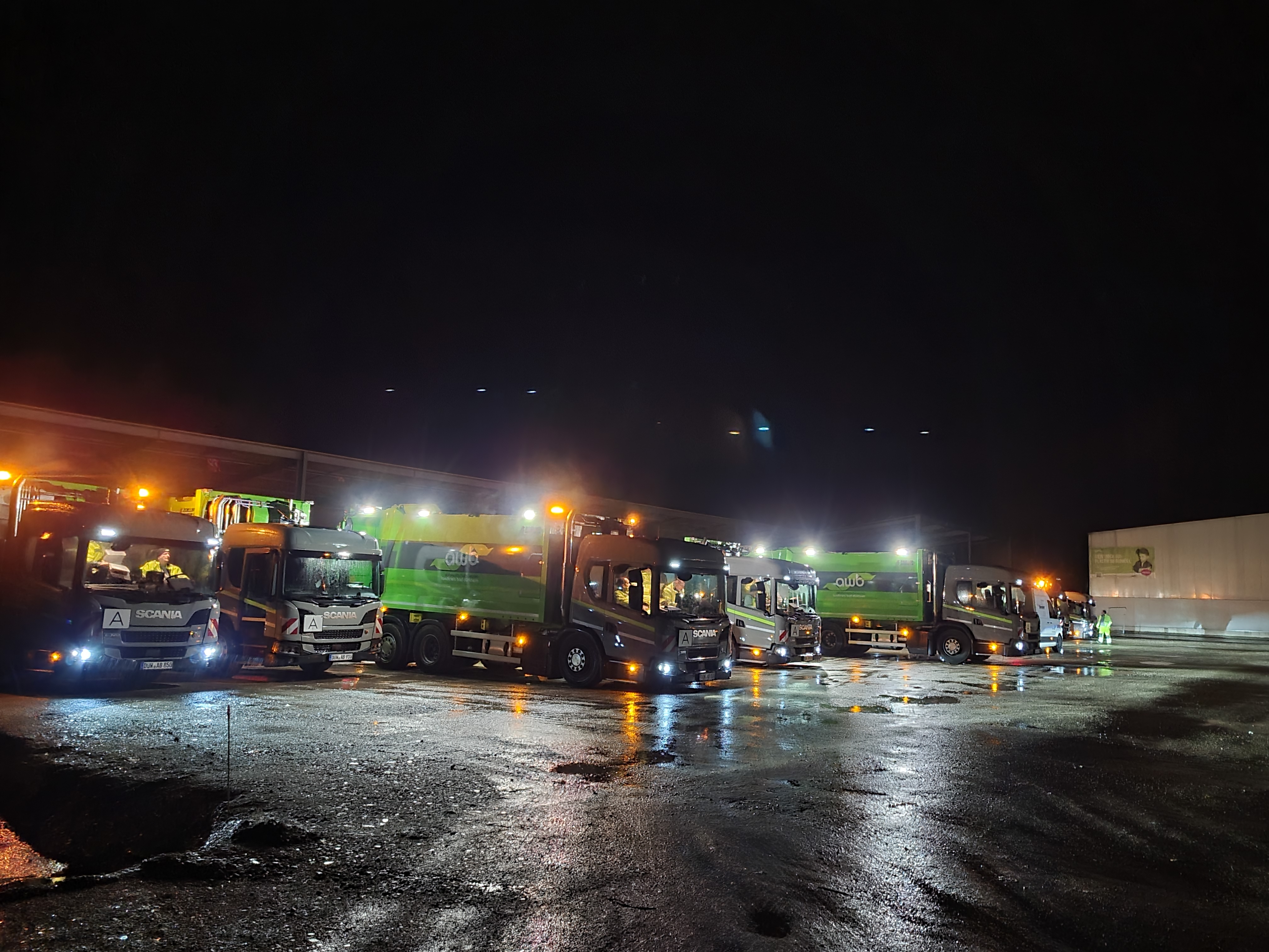 Beleuchtete Abfallsammelfahrzeuge auf einem Betriebsgelände mit Lichtspiegelungen im Regen. 