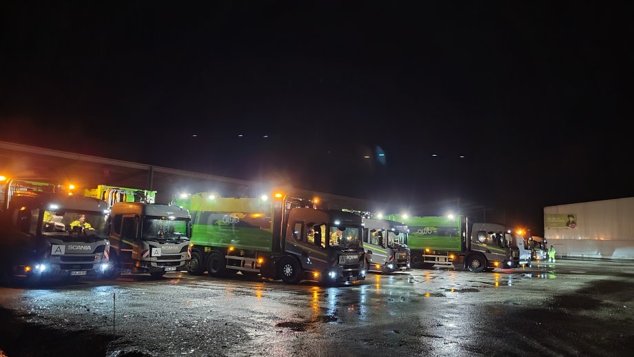 Beleuchtete Abfallsammelfahrzeuge auf einem Betriebsgelände mit Lichtspiegelungen im Regen. 