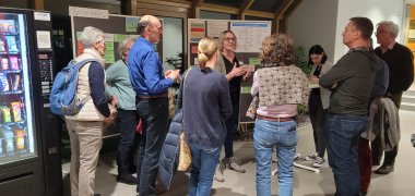 Menschen stehen an einer Mitmachstation bei der Bürgerbeteiligung zum Klimaschutzkonzept und hören einer Frau zu, die gerade spricht. 