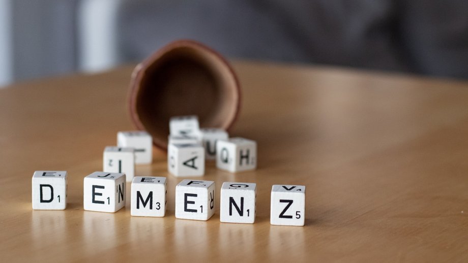 Ein Würfelbecher liegt auf einem Tisch. Davor liegen Buchstaben-Würfel, die da Wort Demenz bilden. 