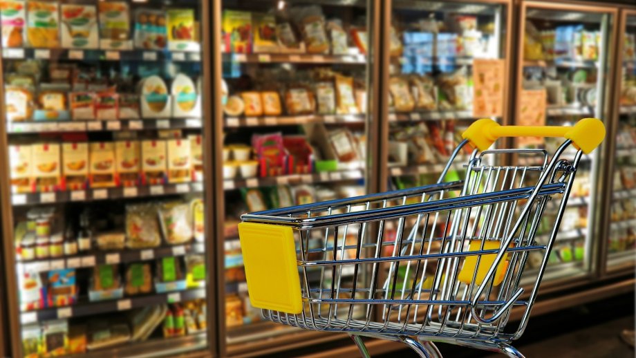 Ein Einkaufswagen steht vor vollen Kühlschränken in einem Supermarkt. 