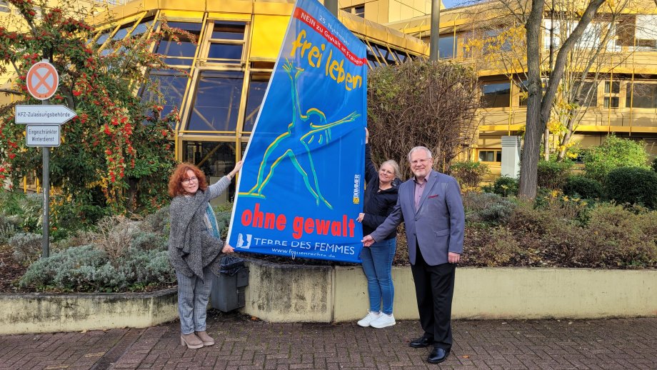 Zwei Frauen und ein Mann halten eine blaue Fahne mit der Aufschrift "Nein zu Gewalt an Frauen, Frei Leben ohne Gewalt". 