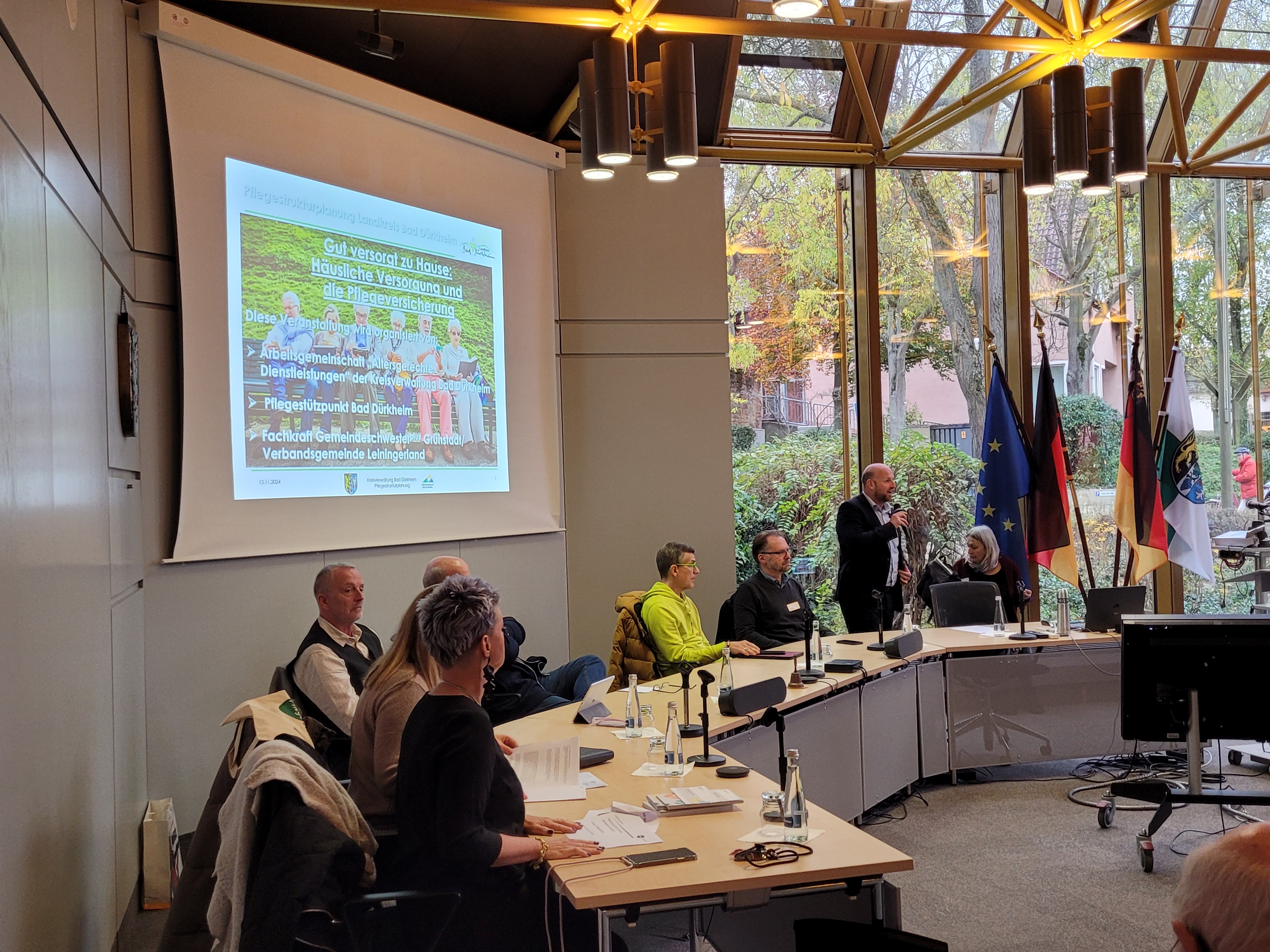 Ein Mann spricht in ein Mikrofon. Daneben sitzen mehrere Menschen. Im Hintergrund eine Präsentation auf einer Leinwand.