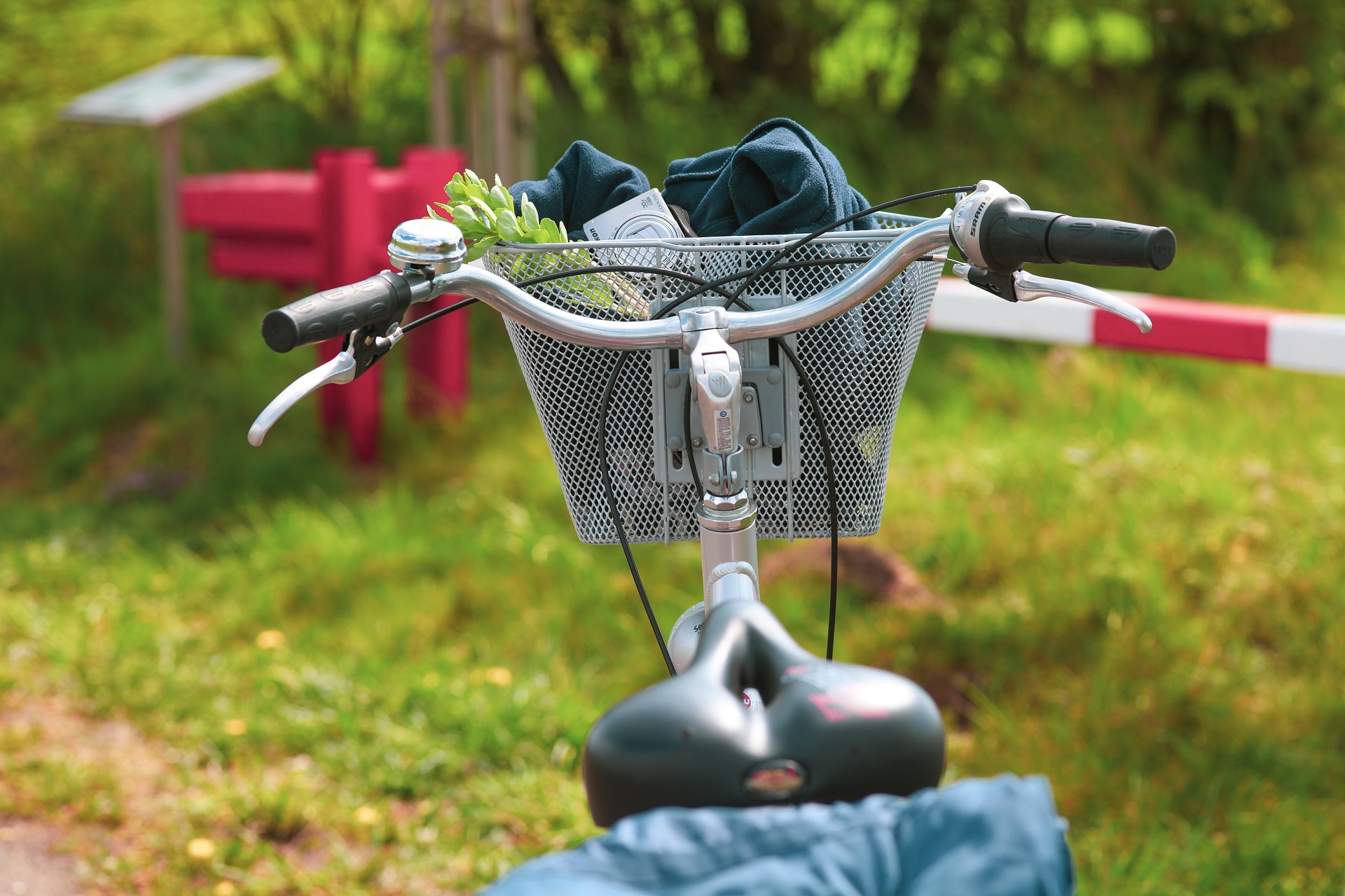 Ein Fahrrad mit einem Korb am Lenker steht im Grünen vor einer rot-weißen Schranke. 