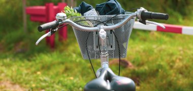 Ein Fahrrad mit einem Korb am Lenker steht im Grünen vor einer rot-weißen Schranke. 