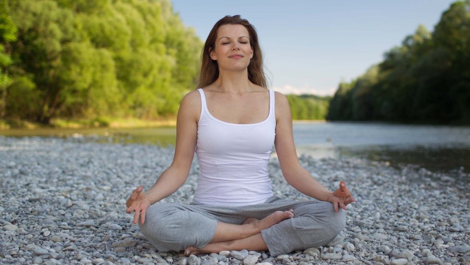 Eine Frau sitzt in Meditationshaltung im Schneidersitz und hat die Augen geschlossen, sie lächelt.
