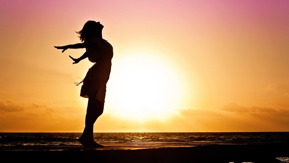 Der Schatten einer Frau mit weit ausgebreiteten Armen vor einem Sonnenuntergang am Meer.