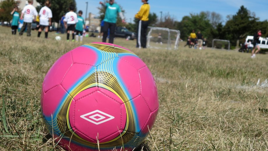 Ein pinkfarbener Fußball liegt auf einem Rasen, im Hintergrund sind Kinder und kleine Tore zu sehen. 