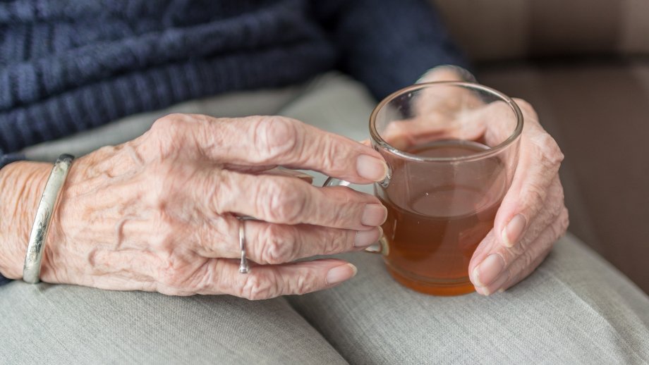 Beide Hände einer älteren Frau, die im Sitzen ein Glas mit Tee hält,