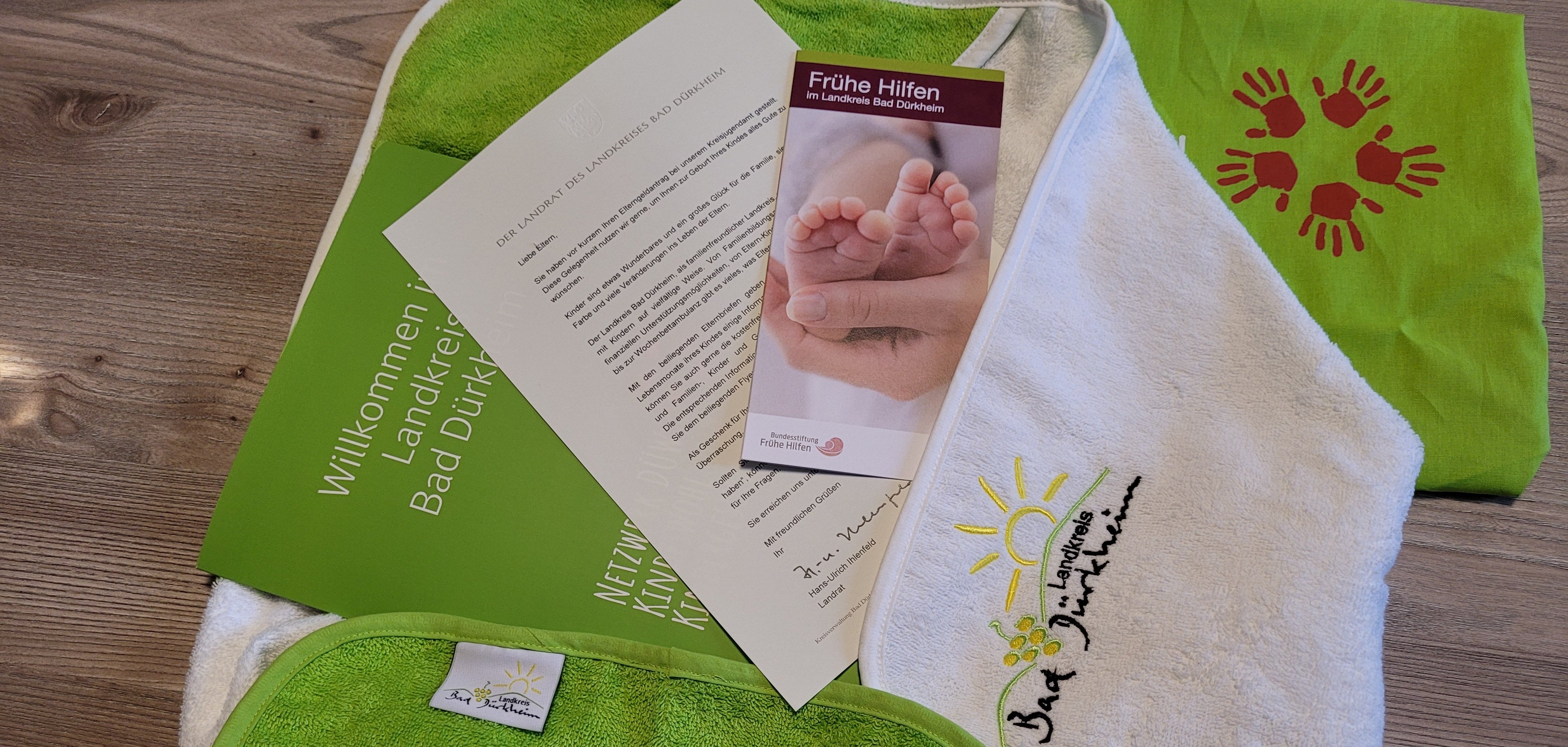 Verschiedene Papiere, eine grüne Tasche mit einem Logo aus roten Handabdrücken, ein weißes Handtuch mit dem Landkreis-Logo und ein grünes Babylätzchen. 
