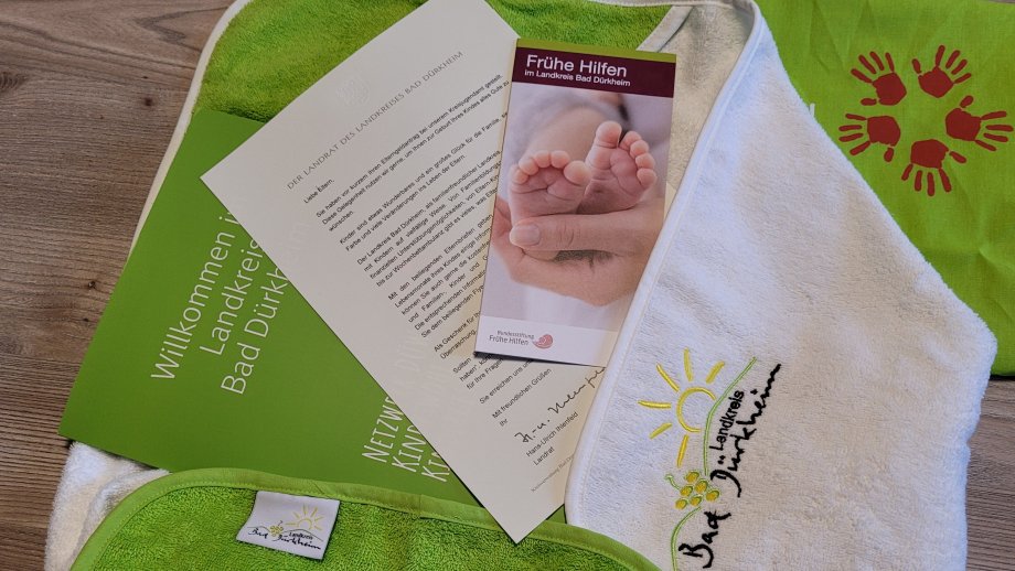 Verschiedene Papiere, eine grüne Tasche mit einem Logo aus roten Handabdrücken, ein weißes Handtuch mit dem Landkreis-Logo und ein grünes Babylätzchen. 