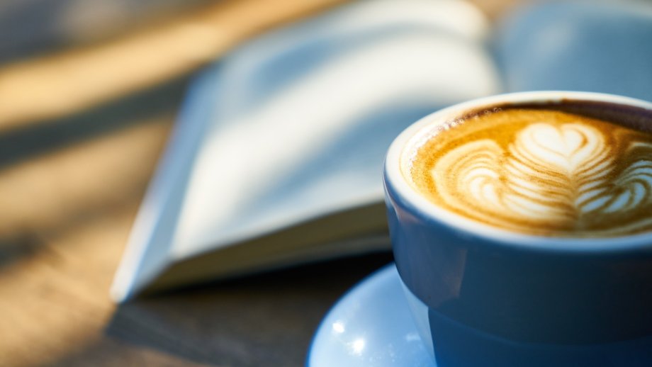 Auf einem Holztisch liegt ein aufgeklapptes Buch, daneben steht eine Tasse Kaffee mit Muster im Milchschaum. 