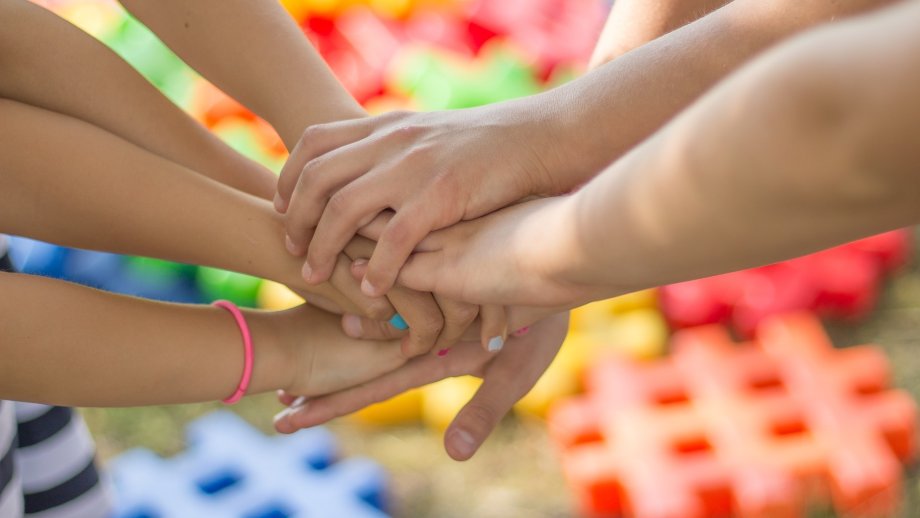 Von im Kreis stehenden Kindern sind nur die in der Kreismitte aufeinander gelegten Hände zu sehen. Im Hintergrund übergroße Schaumstoffpuzzleteile. 