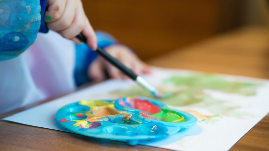 Ein Kind malt mit einem Pinsel auf Papier, im Vordergrund eine bunt verschmierte Palette zum Farbenmischen. 