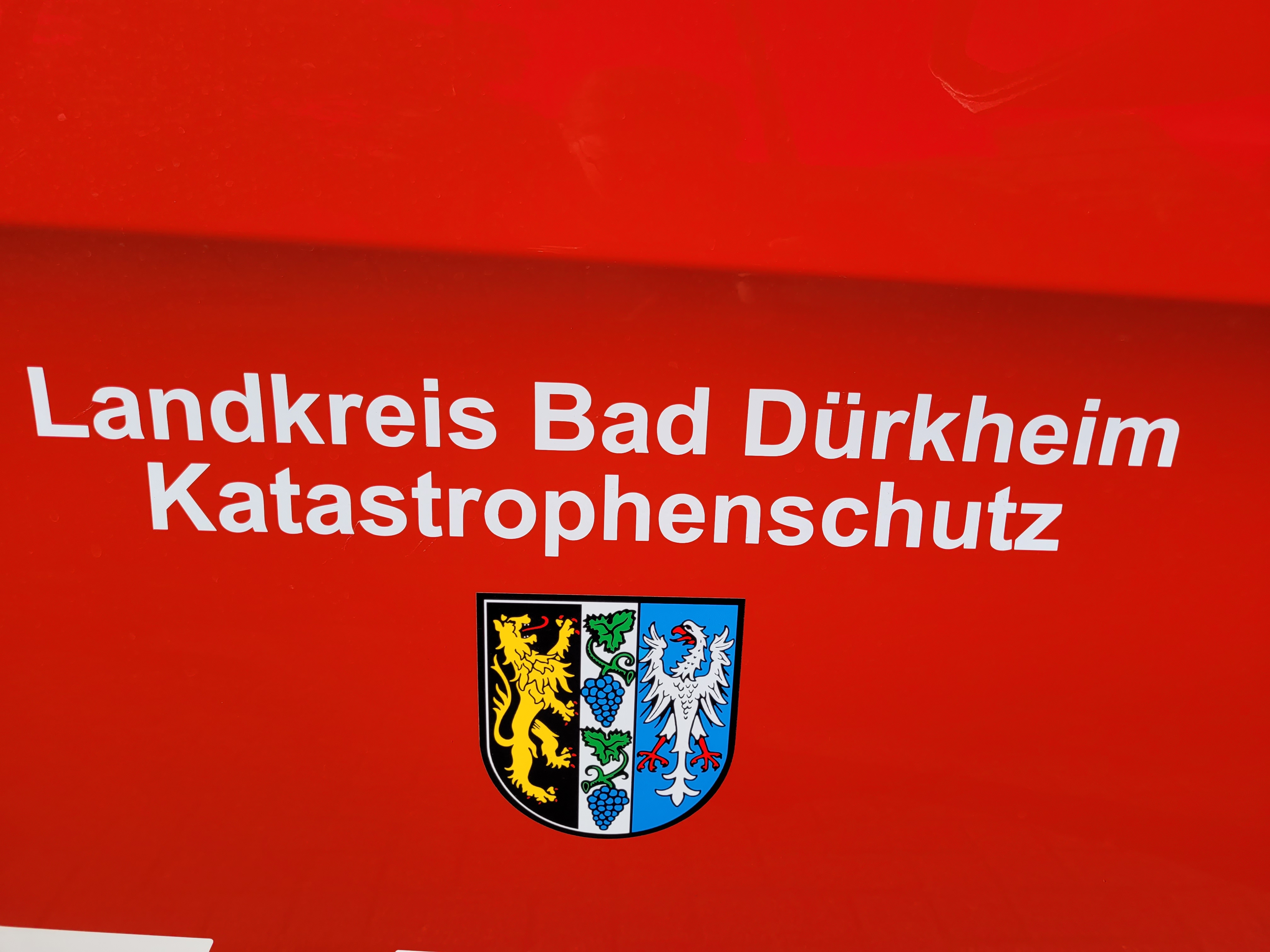 Weißer Schriftzug "Landkreis Bad Dürkheim Katastrophenschutz" über dem Wappen des Landkreises, beides auf feuerwehrrotem Hintergrund. 