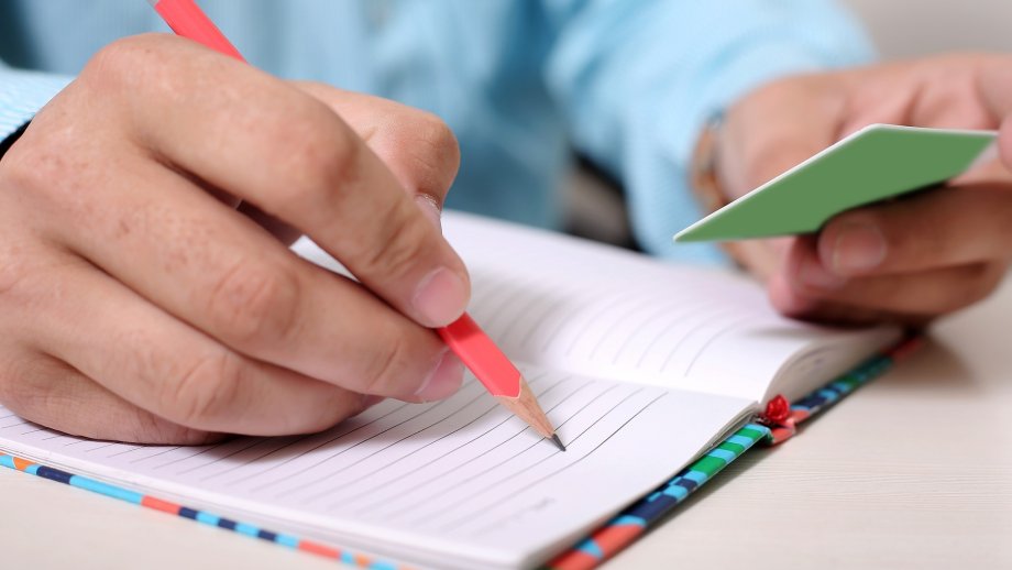 Eine Hand hält einen Bleistift über einem aufgeschlagenen Notizbuch. In der anderen Hand ist die Rückseite einer grünen Karte zu sehen.