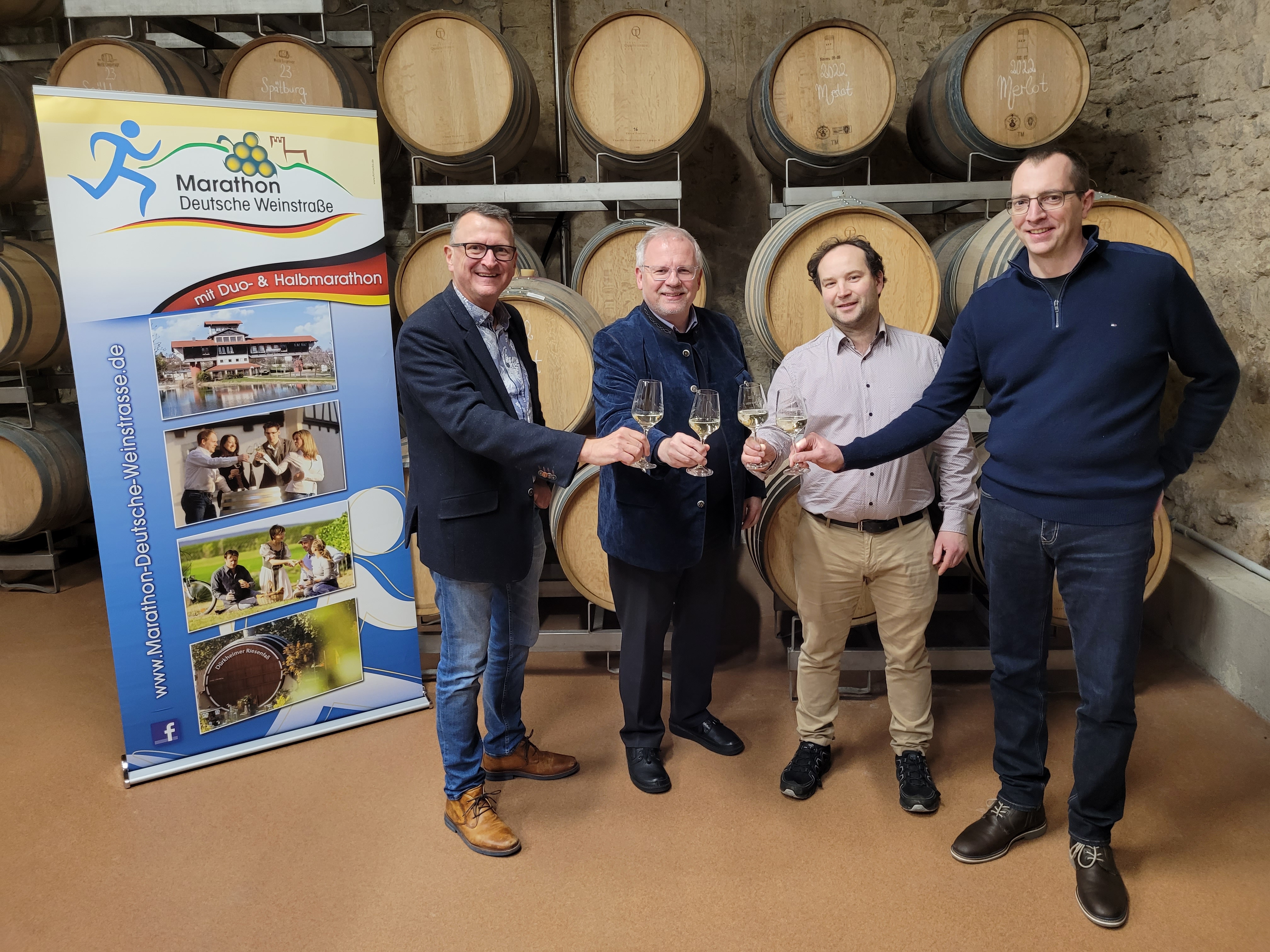 Vier Männer mit einem Glas mit Weißwein in der Hand stehen in einem Keller vor einer Wand mit kleinen Holzfässern. Daneben ein Banner mit Werbung für den Marathon Deutsche Weinstraße. 