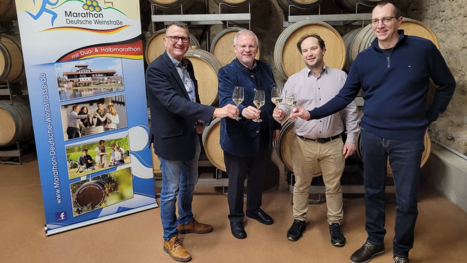 Vier Männer mit einem Glas mit Weißwein in der Hand stehen in einem Keller vor einer Wand mit kleinen Holzfässern. Daneben ein Banner mit Werbung für den Marathon Deutsche Weinstraße. 