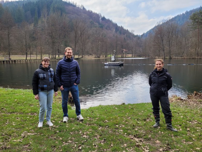 Zwei Frauen und ein Mann stehen am Ufer eines Sees, auf dem ein Plattform mit Geräten schwimmt. 