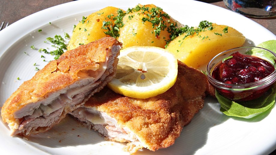 Ein weißer Teller mit einem Cordon Bleu, darauf eine Scheibe Zitrone, daneben Kartoffeln mit Kräutern bestreut und ein Schälchen Preiselbeeren auf einem Salatblatt. 