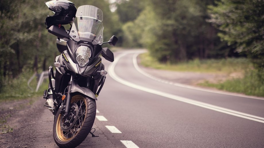 Ein Motorrad steht am Rand einer Landstraße, die eine Kurve macht. Links und rechts sind Bäume zu sehen. 