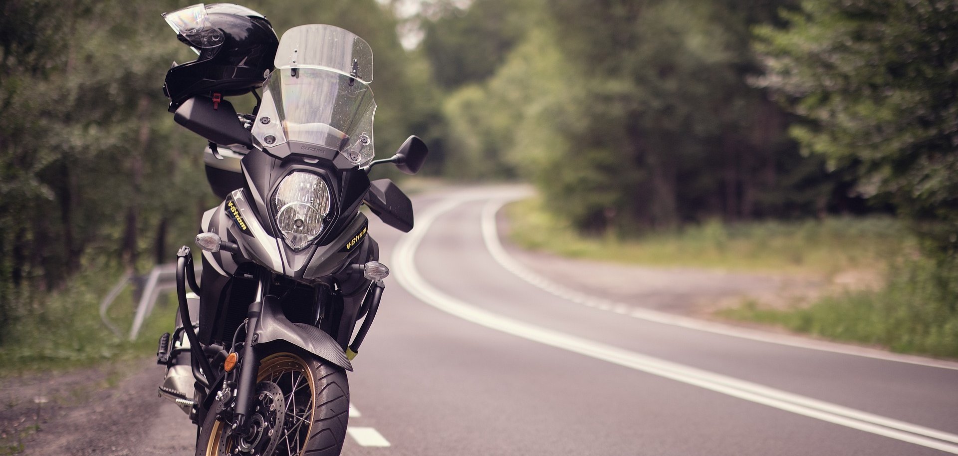 Ein Motorrad steht am Rand einer Landstraße, die eine Kurve macht. Links und rechts sind Bäume zu sehen. 