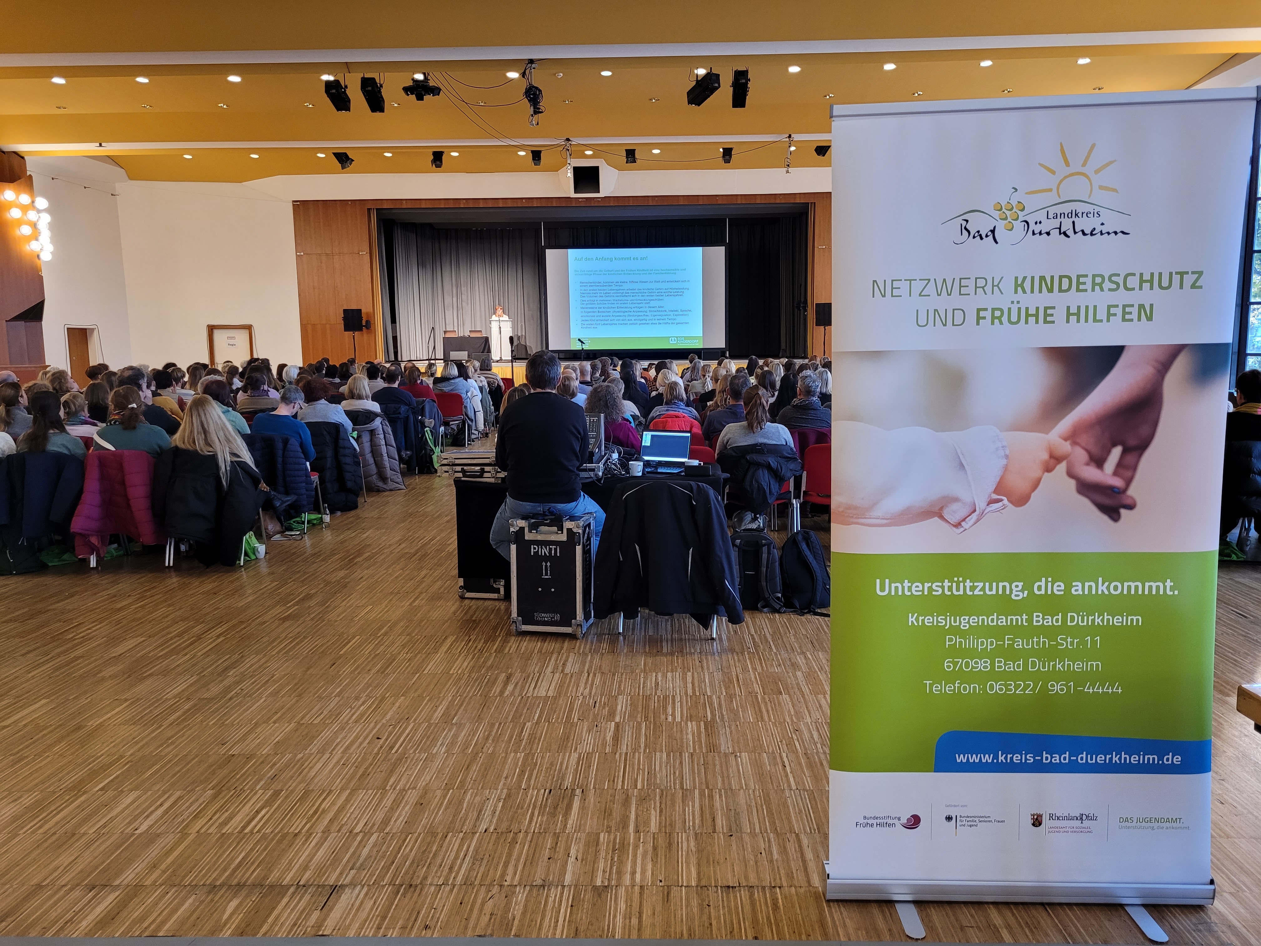 Im Hintergrund eine Bühne, auf der eine Referentin steht. Davor sitzendes Publikum, davor ein Werbe-Roll-up mit Text.