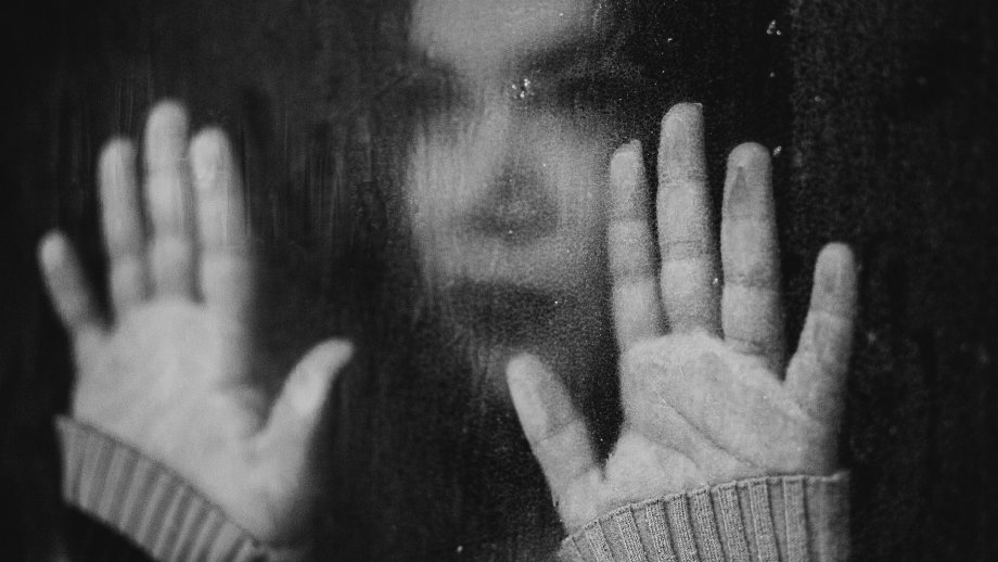 Schwarz-weiß-Foto: Eine traurig aussehende Frau steht hinter einem vom Regen nassen Fenster und hat beide Hände an die Scheibe gelegt. 