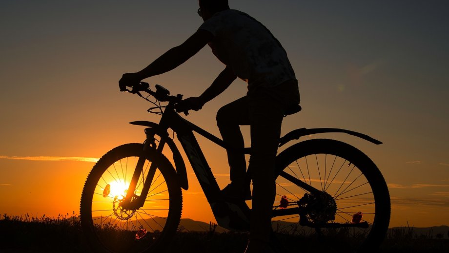 Die Silhouette eines Mannes auf einem E-Bike vor orangefarbenem Sonnenuntergang-Himmel.