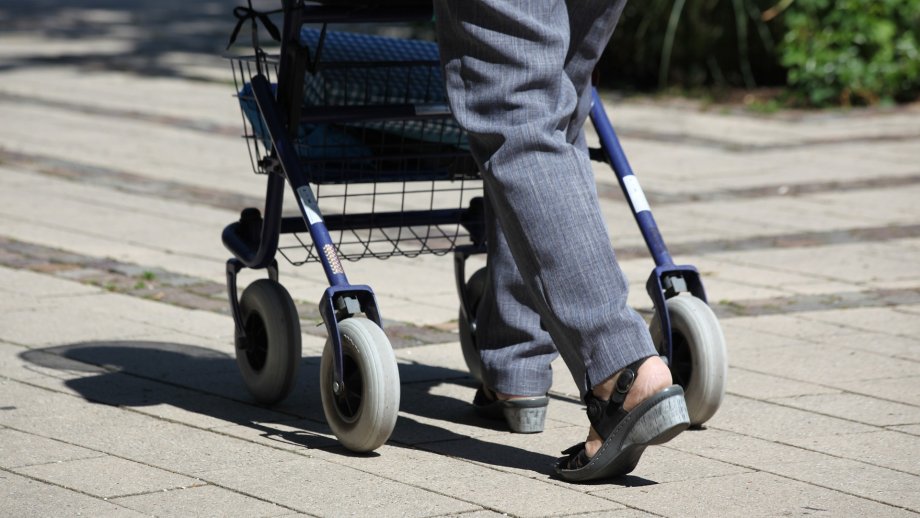 Frau läuft mit Rollator vom Betrachter weg. 