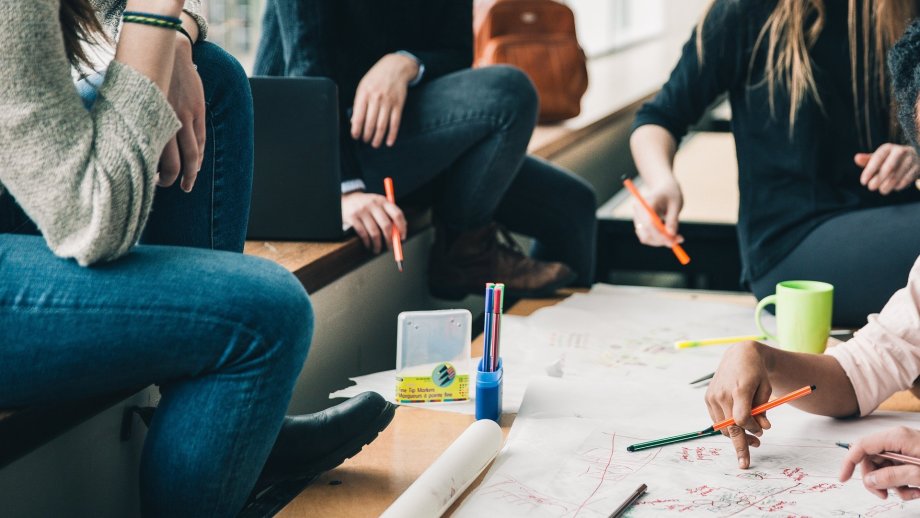 Schülerinnen und Schüler, deren Gesicht nicht zu sehen ist, sitzen um einen Tisch mit einer Gruppenarbeit für die vielen Notizen auf dem Tisch liegen.