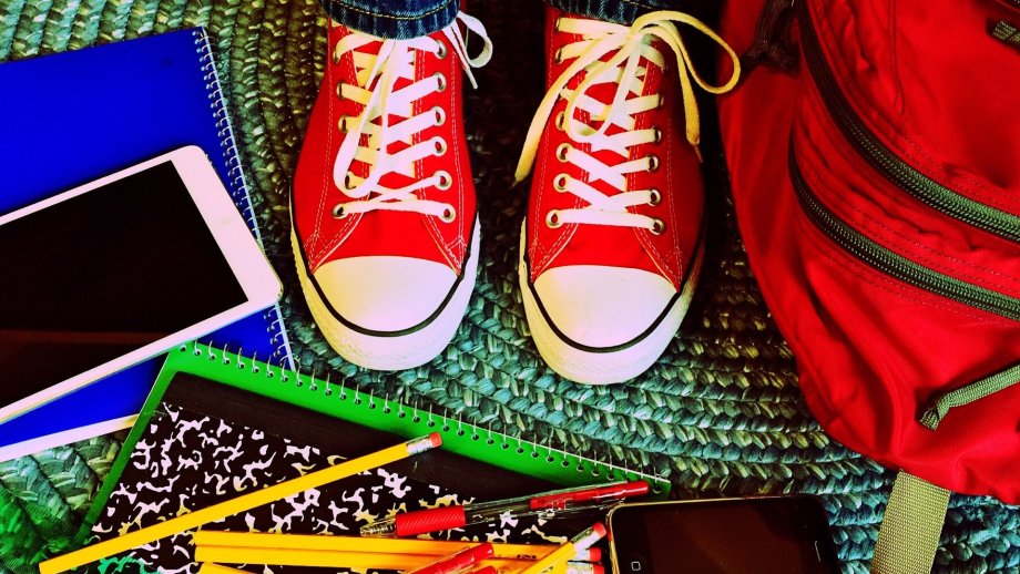 Ein Paar rot-weiße Sneaker, drumherum liegen auf dem Boden ein Smartphone und Schulsachen wie Rucksack, Notizbuch und Stifte. 