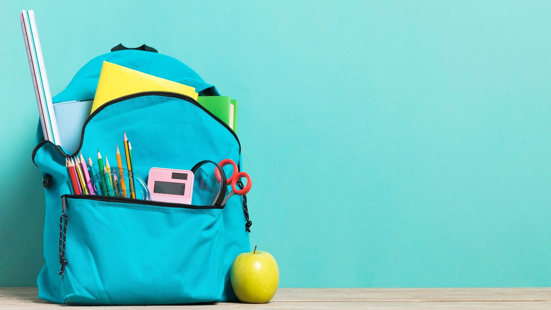 Ein türkisfarbener Rucksack, aus dem Schulmaterialien ragen, steht auf dem Boden vor einer Wand. 