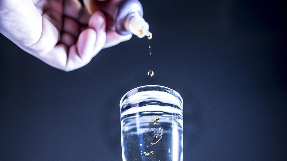 Eine Hand hält ein Fläschchen, aus dem Tropfen in ein Glas fallen.