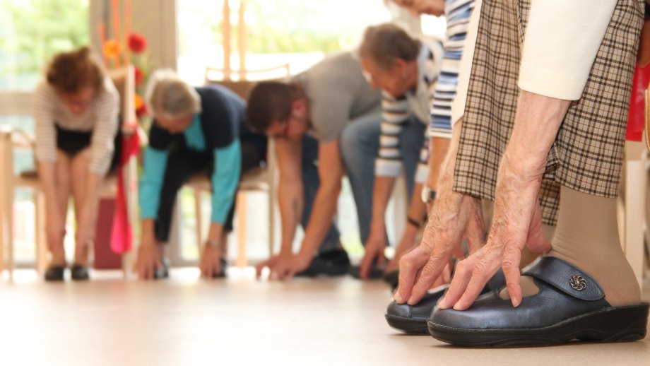 Seniorinnen und Senioren sitzen auf Stühlen in einer Reihe und versuchen, mit ihren Händen die Füße zu erreichen. 