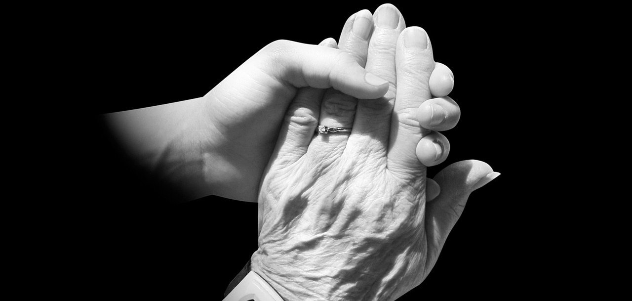 Eine junge Hand hält eine alte Hand mit einem Ring am Ringfinger und einem Notrufknopf am Handgelenk. 
