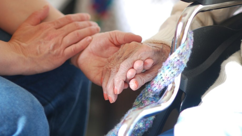 Zwei junge Hände halten eine Hand einer älteren Frau mit zartrosa lackierten Fingernägeln. 