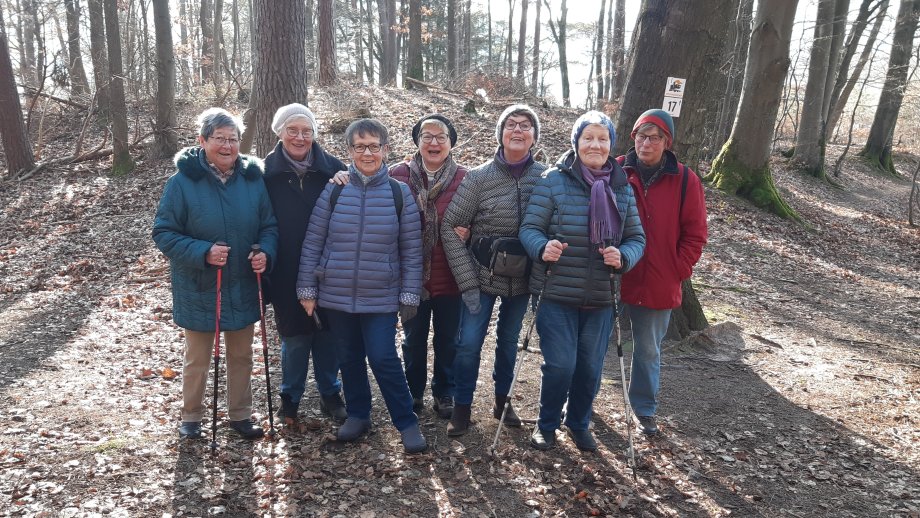 Eine Gruppe Seniorinnen steht in Herbstkleidung im Wald auf laubbedecktem Boden. 