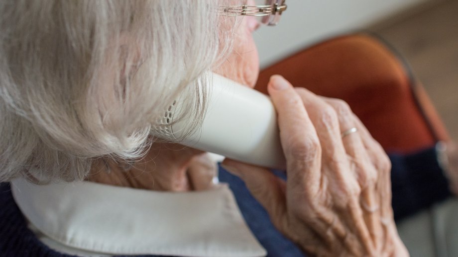 Eine Seniorin mit Brille, deren Gesicht nur seitlich undeutlich zu sehen ist, hält einen weißen Telefonhörer ans Ohr. 