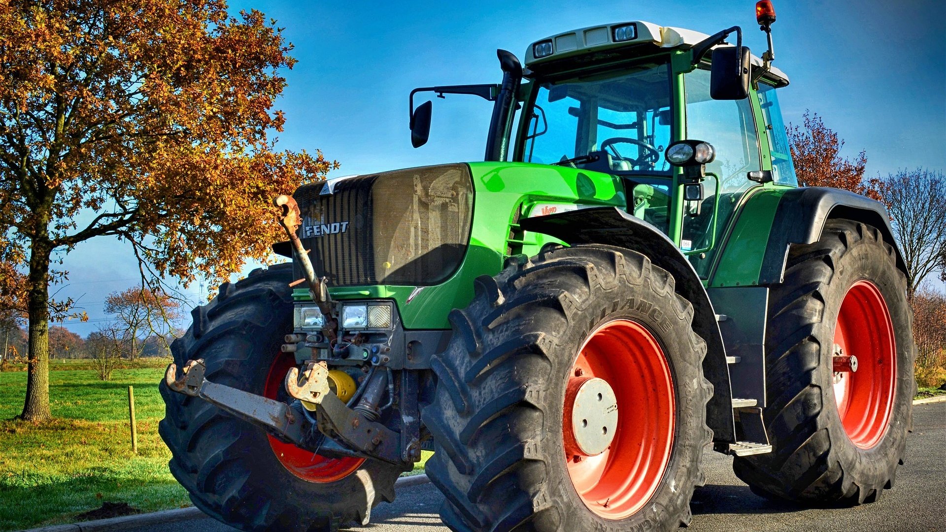 Ein großer Traktor auf einer Straße, im Hintergrund ein Baum mit Herbstlaub auf einer Wiese vor blauem Himmel. 